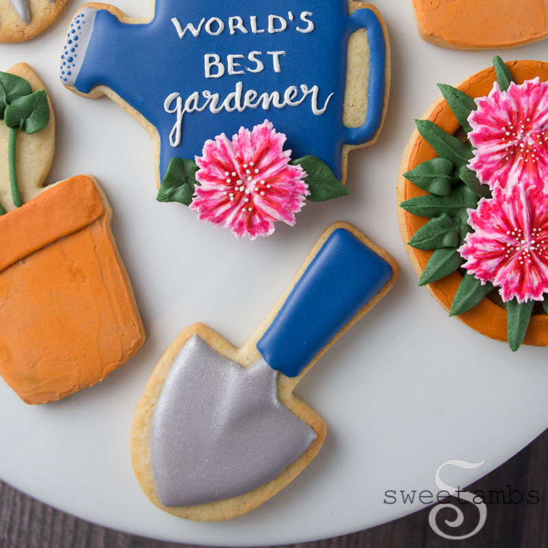 A cookie decorated to look like a hand shovel. At the top of the photo is a cookie decorated to look like a watering can with a dianthus on it and the words "World's Best Gardener" written in icing. 