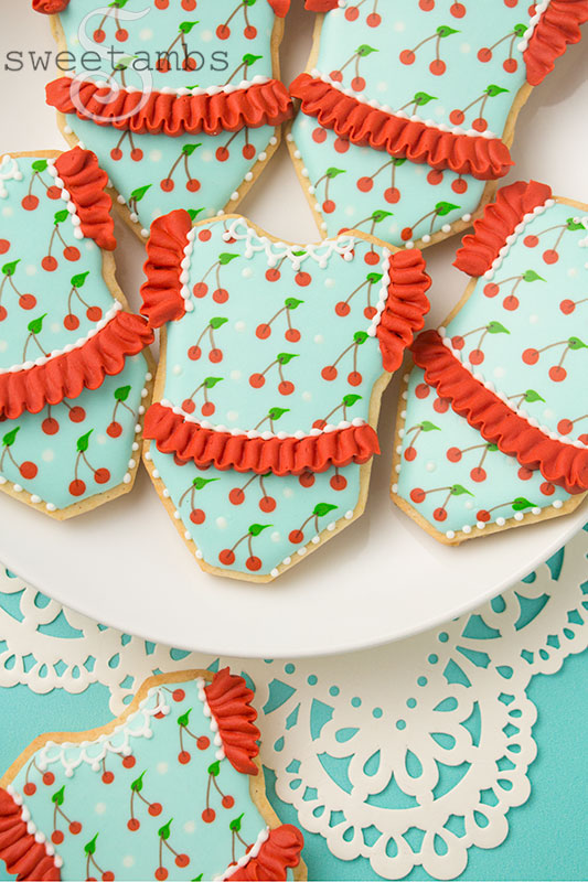 Cookies decorated to look like baby rompers with a cherry pattern. The cookies have icing ruffles on the sleeves and in the middle, as well as lace detail along the color. The cookies are sitting on a plate over a lace doily.