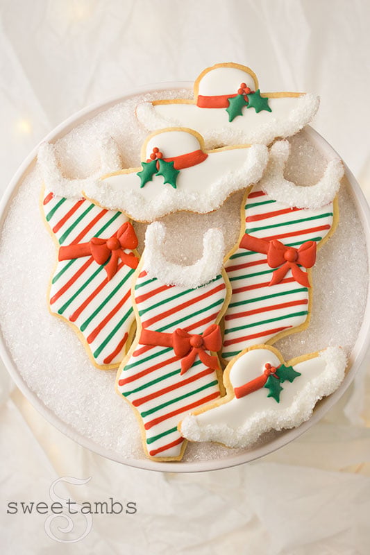 A set of cookies decorated to look like bathing suits and sun hats with holy leaves and berries, candy cane stripes, and gift bows. The collar of the bathing suit cookies and the edge of the sun hat cookies are trimmed with sparkling sugar. The cookies are on a platter covered with sparkling sugar and under the platter are soft fairy lights.