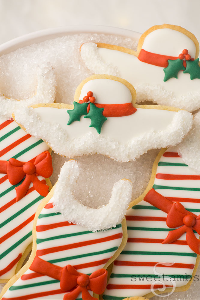 A close up of cookies decorated to look like bathing suits and sun hats with holy leaves and berries, candy cane stripes, and gift bows. The collar of the bathing suit cookies and the edge of the sun hat cookies are trimmed with sparkling sugar. The cookies are on a platter covered with sparkling sugar. There are soft fairy lights in the background.