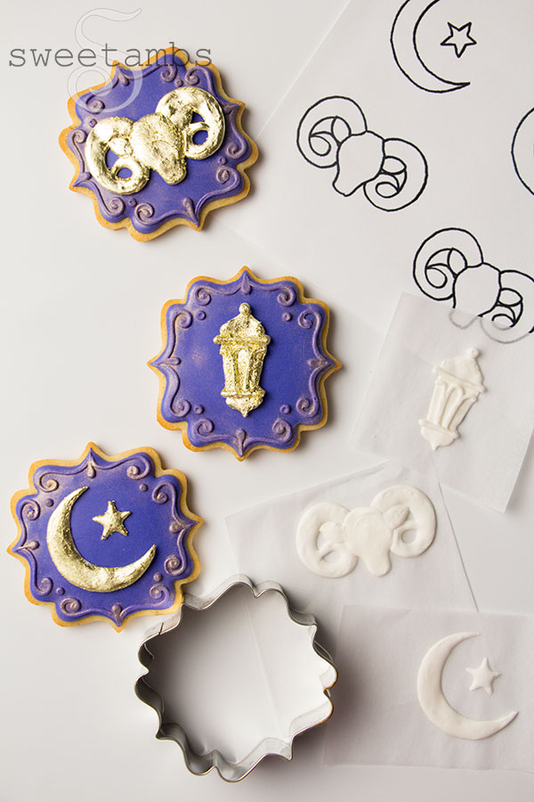 Ornate plaque shaped cookies decorated with deep purple icing. The cookies are adorned with a gold lantern, a gold crescent moon and star, and a gold ram. The edges of the cookie are decorated with a filigree design and gold luster dust. Next to the cookies is the Eid symbol template, 3 royal icing transfers on squares of parchment paper in the shape of Eid symbols, and a square plaque cookie cutter.