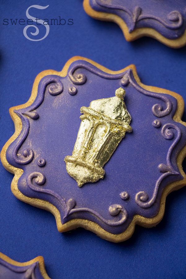 Ornate plaque shaped cookies decorated with deep purple icing. The cookie is adorned with a gold lantern. The edges of the cookie are decorated with a filigree design and gold luster dust.