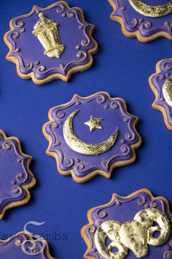 Ornate plaque shaped cookies decorated with deep purple icing. The cookies are adorned with a gold lantern, a gold crescent moon and star, and a gold ram. The edges of the cookie are decorated with a filigree design and gold luster dust.