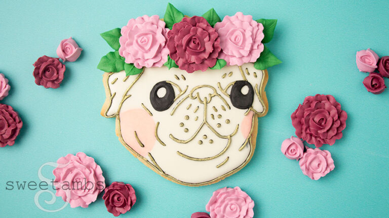 A cookie decorated to look like a pug face. The outlines on the face are painted with gold and the cheeks are tinted with peachy pink. The pug has 3 royal icing flowers with leaves as a crown. The cookie is surrounded by royal icing flowers.