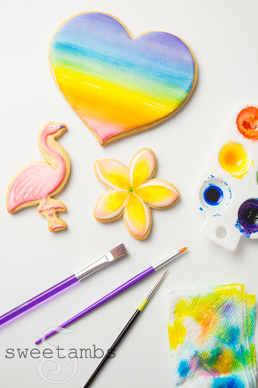 A heart cookie painted with a rainbow watercolor wash, a flamingo cookie, and a plumeria cookie. The cookies are surrounded by a paint palette with food coloring watercolors, paint brushes, and a paper towel used for blotting the brushes.