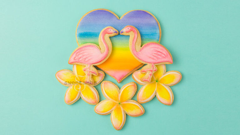 A set of cookies decorated to look like a tropical scene containing a rainbow watercolor heart, two flamingos facing each other, and three plumeria underneath the heart and the flamingos.