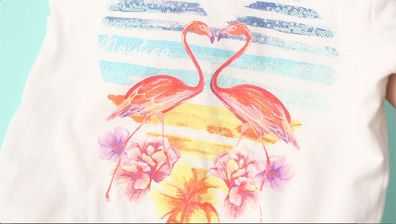 A t-shirt with a tropical scene containing a heart with a sunset, two flamingos facing each other, and a variety of tropical flowers underneath the heart and the flamingos.