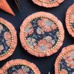 A set of round cookies decorated with peach and pink flowers on a dark blue background. There is a royal icing ruffled edge around the cookies. In between the cookies are piping bags filled with royal icing.