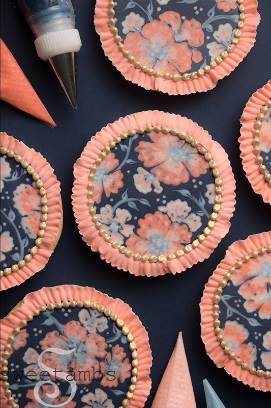 A set of round cookies decorated with peach and pink flowers on a dark blue background. There is a royal icing ruffled edge around the cookies. In between the cookies are piping bags filled with royal icing.