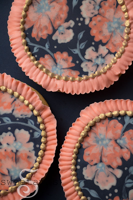 A set of round cookies decorated with peach and pink flowers on a dark blue background. There is a royal icing ruffled edge around the cookies and a gold bead border along the inside edge of the ruffle.