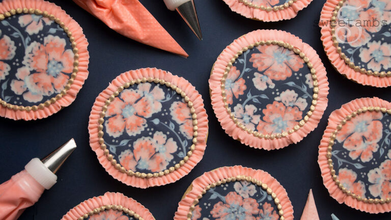 A set of round cookies decorated with peach and pink flowers on a dark blue background. There is a royal icing ruffled edge around the cookies. In between the cookies are piping bags filled with royal icing.