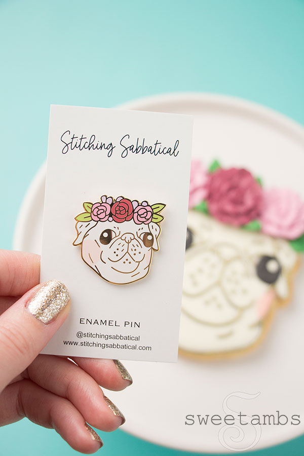 An enamel pin of a pug face. The outlines on the face are gold and the cheeks are tinted with peachy pink. The pug has 3 flowers with leaves as a crown. There is a pug cookie that matches the pin in the background. The pin is being held by a thumb and fingers with glitter nail polish