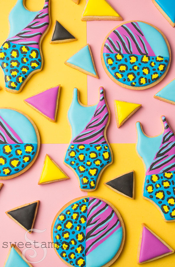 A set of cookies decorated with brightly colored animal prints. There are bathing suit cookies, round cookies, and smaller triangle cookies in solid colors. The cookies are on top of a color block background.