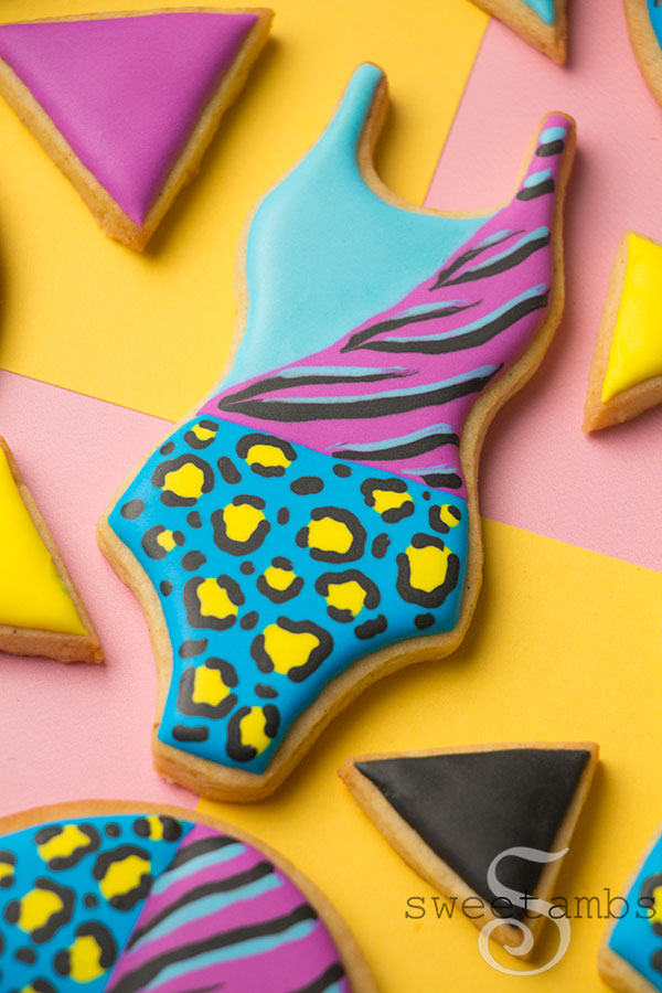 A bathing suit cookie decorated with brightly colored animal prints and smaller triangle cookies in solid colors. The cookies are on top of a color block background. 