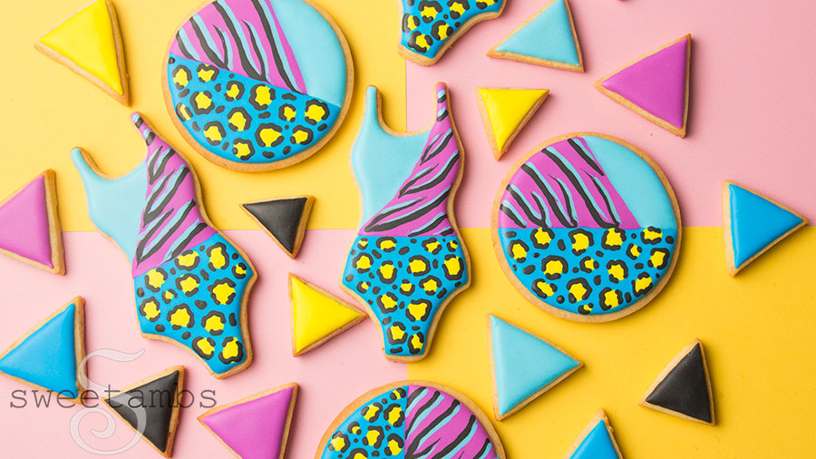 A set of cookies decorated with brightly colored animal prints. There are bathing suit cookies, round cookies, and smaller triangle cookies in solid colors. The cookies are on top of a color block background. 