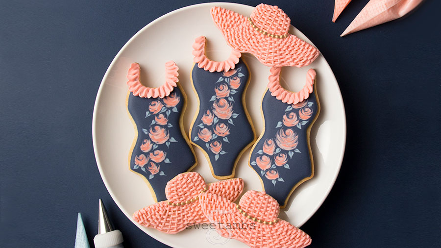 A set of bathing suit cookies decorated with wet-on-wet royal icing roses and a ruffle along the neckline and sunhat cookies decorated with a basket weave pattern and a gold beaded band. The cookies are on a plate on a solid color surface. There are decorating bags filled with icing next to the plate of cookies.