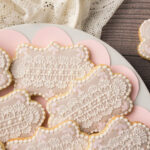 A set of brush embroidered cookies decorated with a royal icing lace design. There are dots of gold on the cookies and the cookies are edged with a bead border. The cookies are sitting on a plate lined with a scalloped liner. There is an antique style lace fabric in the background.