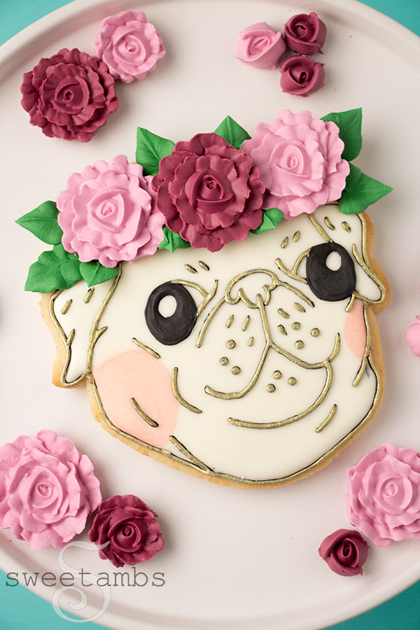 A pug cookie decorated to look like a pug face. The outlines on the face are painted with gold and the cheeks are tinted with peachy pink. The pug has 3 royal icing flowers with leaves as a crown. The cookie is surrounded by royal icing flowers.