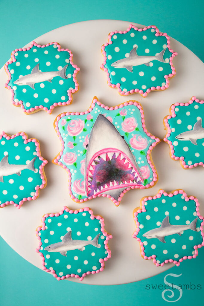 Plaque shaped cookies decorated with roses, polka dots, and sharks. The cookie in the middle is decorated with a shark with an open mouth showing its teeth. The smaller cookies surrounding it are decorated with profiles of small sharks.
