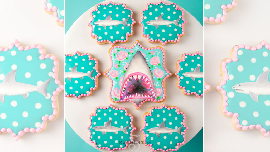 Plaque shaped cookies decorated with roses, polka dots, and sharks. The cookie in the middle is decorated with a shark with an open mouth showing its teeth. The smaller cookies surrounding it are decorated with profiles of small sharks.