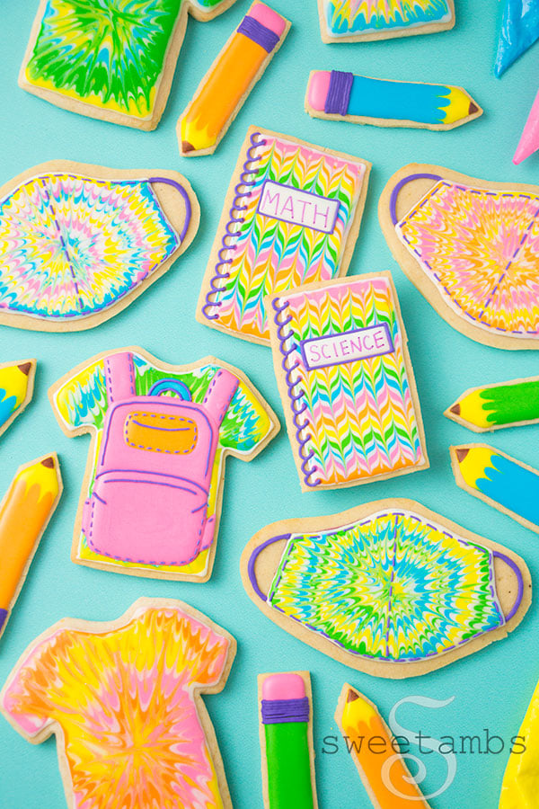 Back to school cookies. This set of cookies is decorated with brightly colored royal icing to look like tie dye t-shirts, a backpack, pencils, spiral notebooks with the words Math and Science written on them, and tie dye masks. There are tipless decorating bags filled with icing around the cookies in the upper right corner of the image.