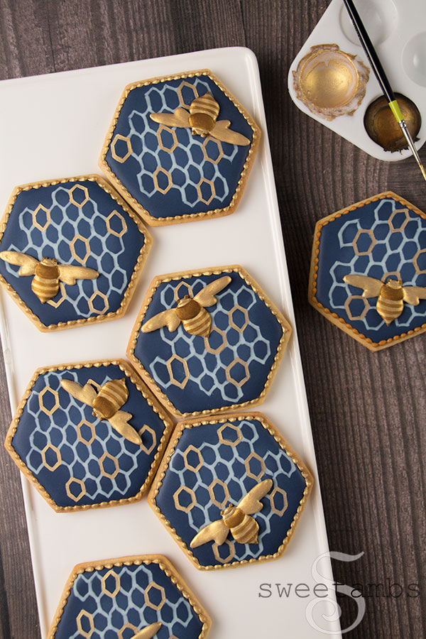 Decorated bee cookies. The cookies are hexagon shaped with navy blue royal icing and a light blue and gold honeycomb pattern. There are royal icing bees on the cookies painted with gold and bronze edible luster dust. The cookies have a gold bead border around the edges. The cookies are on a white rectangle pattern on a wooden surface. In the upper righthand corner there is a paint palette with gold and bronze edible paint with a thin brush lying across it.