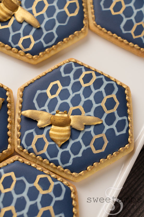 A close up of decorated bee cookies. The cookies are hexagon shaped with navy blue royal icing and a light blue and gold honeycomb pattern. There are royal icing bees on the cookies painted with gold and bronze edible luster dust. The cookies have a gold bead border around the edges. The cookies are on a white rectangle pattern on a wooden surface.