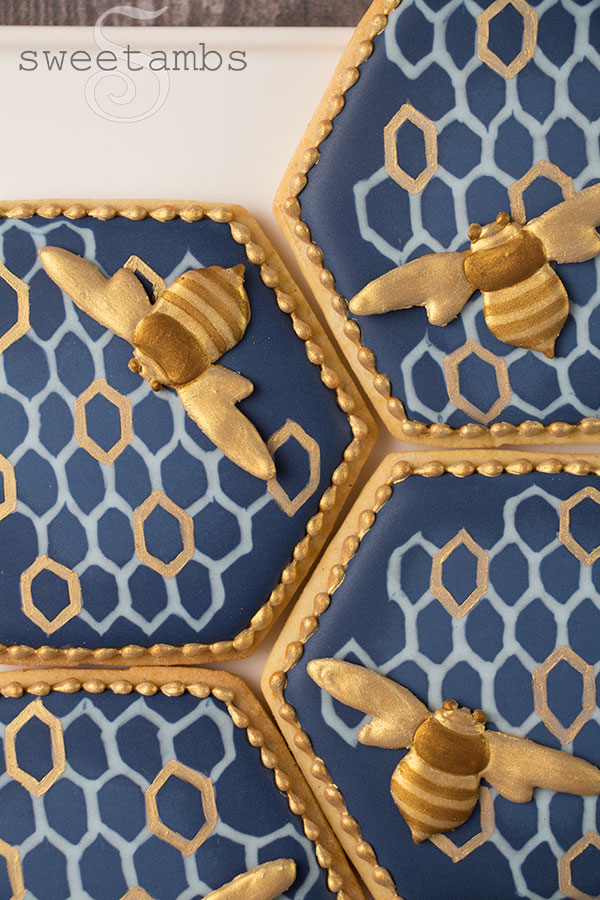 A close up of decorated bee cookies. The cookies are hexagon shaped with navy blue royal icing and a light blue and gold honeycomb pattern. There are royal icing bees on the cookies painted with gold and bronze edible luster dust. The cookies have a gold bead border around the edges. The cookies are on a white rectangle pattern on a wooden surface.