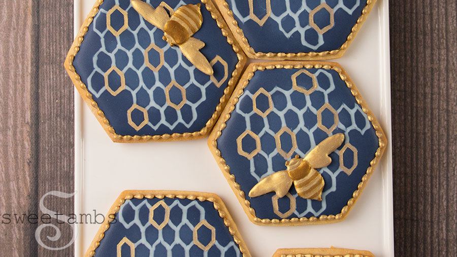 Decorated bee cookies. The cookies are hexagon shaped with navy blue royal icing and a light blue and gold honeycomb pattern. There are royal icing bees on the cookies painted with gold and bronze edible luster dust. The cookies have a gold bead border around the edges. The cookies are on a white rectangle pattern on a wooden surface.