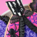 A close-up of the Spider Queen Halloween Cookie. This cookie is in the shape of a gown. The cookie is decorated with black and white stripes and a spider on the bodice. The top layer of the skirt is hot pink with purple spider webs and a spider on each side. The edge of the top layer is decorated with black ruffles. There is brush embroidery lace layered on the top of the skirt. The bottom layer of the skirt is purple with light purple filigree. The top of the bodice has a black bead border. The cookie is on a platter covered with silver sparkling sugar over a pink backdrop.