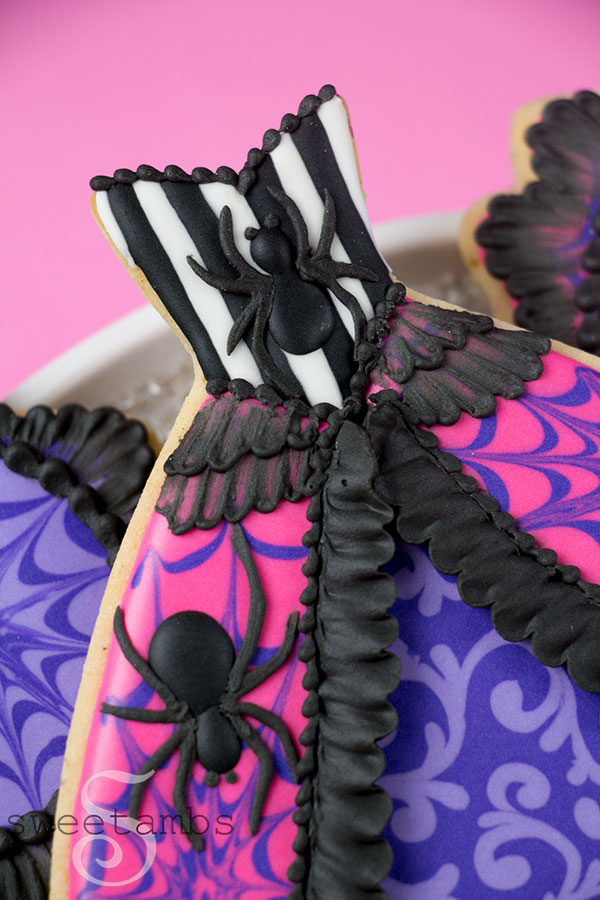A close-up of the Spider Queen Halloween Cookie. Spider Queen Halloween Cookie - a cookie in the shape of a gown. The cookie is decorated with black and white stripes and a spider on the bodice. The top layer of the skirt is hot pink with purple spider webs and a spider on each side. The edge of the top layer is decorated with black ruffles. There is brush embroidery lace layered on the top of the skirt. The bottom layer of the skirt is purple with light purple filigree. The top of the bodice has a black bead border. The cookie is on a platter covered with silver sparkling sugar over a pink backdrop.