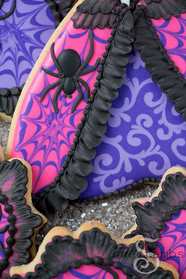 A close-up of the skirt on the Spider Queen Cookie. This cookie is in the shape of a gown. The cookie is decorated with black and white stripes and a spider on the bodice. The top layer of the skirt is hot pink with purple spider webs and a spider on each side. The edge of the top layer is decorated with black ruffles. There is brush embroidery lace layered on the top of the skirt. The bottom layer of the skirt is purple with light purple filigree. 