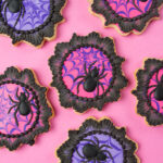 Spider cookies for Halloween - These cookies are cut into a plaque shape and decorated with spider webs in hot pink and purple. There are black royal icing spiders on the cookies and the cookies are finished with a black brush embroidered lace border. The cookies are on a pink background.