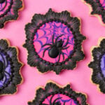 Spider cookies for Halloween - These cookies are cut into a plaque shape and decorated with spider webs in hot pink and purple. There are black royal icing spiders on the cookies and the cookies are finished with a black brush embroidered lace border. The cookies are on a pink background.