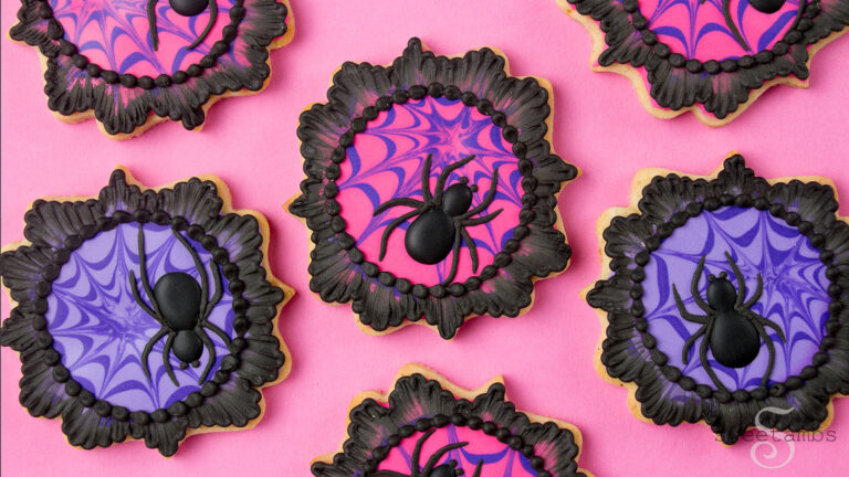 Spider cookies for Halloween - These cookies are cut into a plaque shape and decorated with spider webs in hot pink and purple. There are black royal icing spiders on the cookies and the cookies are finished with a black brush embroidered lace border. The cookies are on a pink background.