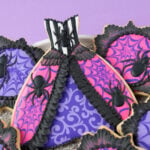 Spider Queen Halloween Cookie - a cookie in the shape of a gown. The cookie is decorated with black and white stripes and a spider on the bodice. The top layer of the skirt is hot pink with purple spider webs and a spider on each side. The edge of the top layer is decorated with black ruffles. There is brush embroidery lace layered on the top of the skirt. The bottom layer of the skirt is purple with light purple filigree. The bottom of the skirt has a black bead border. The top of the bodice also has a black bead border. The gown cookie is surrounded by small plaque shaped cookies decorated with a black brush embroidery border, bright pink and purple icing, and purple spider webs. The plaque cookies have black royal icing spiders on them. The cookies are on a platter covered with silver sparkling sugar over a purple backdrop.