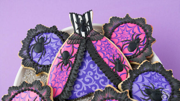Spider Queen Halloween Cookie - a cookie in the shape of a gown. The cookie is decorated with black and white stripes and a spider on the bodice. The top layer of the skirt is hot pink with purple spider webs and a spider on each side. The edge of the top layer is decorated with black ruffles. There is brush embroidery lace layered on the top of the skirt. The bottom layer of the skirt is purple with light purple filigree. The bottom of the skirt has a black bead border. The top of the bodice also has a black bead border. The gown cookie is surrounded by small plaque shaped cookies decorated with a black brush embroidery border, bright pink and purple icing, and purple spider webs. The plaque cookies have black royal icing spiders on them. The cookies are on a platter covered with silver sparkling sugar over a purple backdrop.