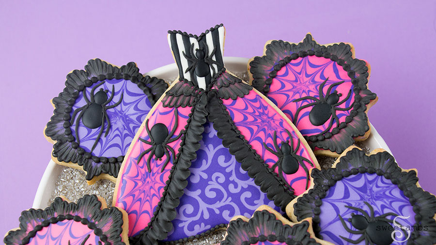 Spider Queen Halloween Cookie - a cookie in the shape of a gown. The cookie is decorated with black and white stripes and a spider on the bodice. The top layer of the skirt is hot pink with purple spider webs and a spider on each side. The edge of the top layer is decorated with black ruffles. There is brush embroidery lace layered on the top of the skirt. The bottom layer of the skirt is purple with light purple filigree. The bottom of the skirt has a black bead border. The top of the bodice also has a black bead border. The gown cookie is surrounded by small plaque shaped cookies decorated with a black brush embroidery border, bright pink and purple icing, and purple spider webs. The plaque cookies have black royal icing spiders on them. The cookies are on a platter covered with silver sparkling sugar over a purple backdrop. 