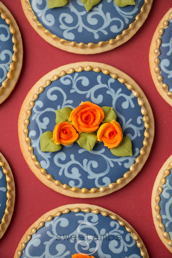 Fall cookies decorated with navy blue icing and a light blue filigree design. The cookies are decorated with orange royal icing roses with green leaves. There is a gold bead border around the edge of the cookies. The cookies are on a burgundy background.