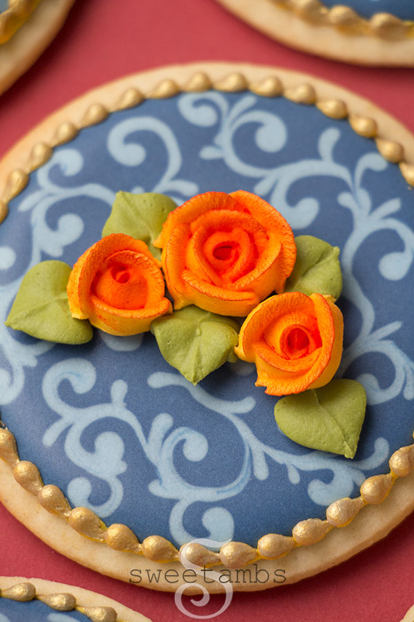 A close up of Fall cookies decorated with navy blue icing and a light blue filigree design. The cookies are decorated with orange royal icing roses with green leaves. There is a gold bead border around the edge of the cookies. The cookies are on a burgundy background.