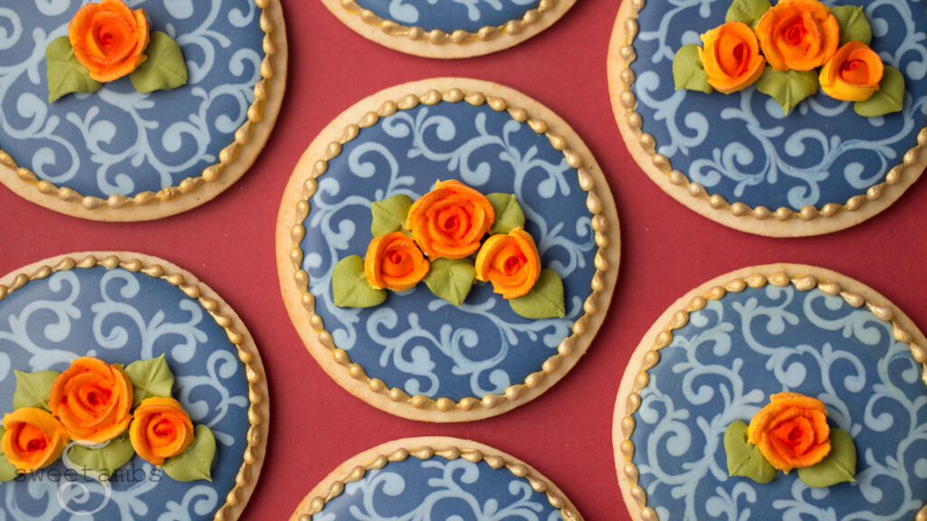 Fall cookies decorated with navy blue icing and a light blue filigree design. The cookies are decorated with orange royal icing roses with green leaves. There is a gold bead border around the edge of the cookies. The cookies are on a burgundy background.