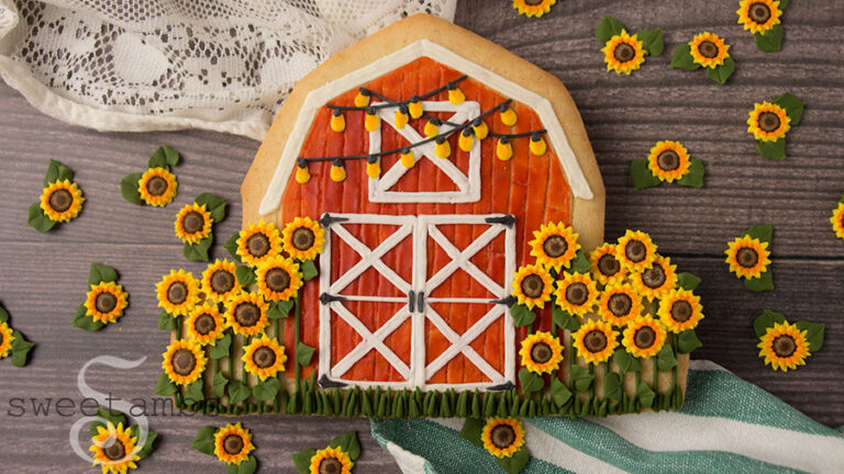 Royal icing sunflowers and barn cookie - A large cookie shaped and decorated to look like a red barn with a white roof and white boards across the window and doors. There are royal icing string lights across the top of the barn cookie. The cookie is surrounded by royal icing sunflowers. The cookie and sunflowers are on a wood background. There is a green and white striped fabric below the cookie and a white lace fabric above the cookie.