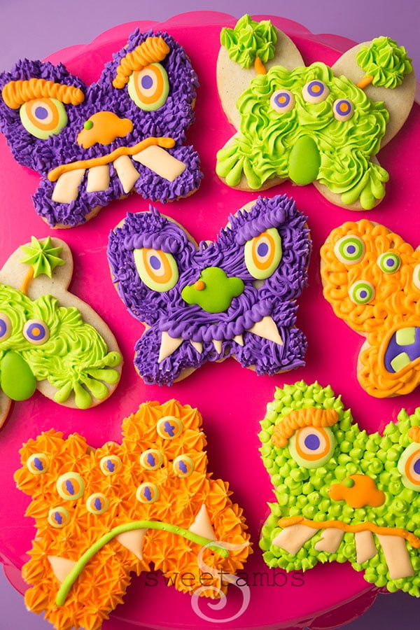 Monster Cookies for Halloween - these cookies are cut from a butterfly cookie cutter and decorated to look like monster faces. The monsters are decorated in shades of green, orange, and purple royal icing with lots of different textures. The cookies are on a bright pink cake plate on a purple background.