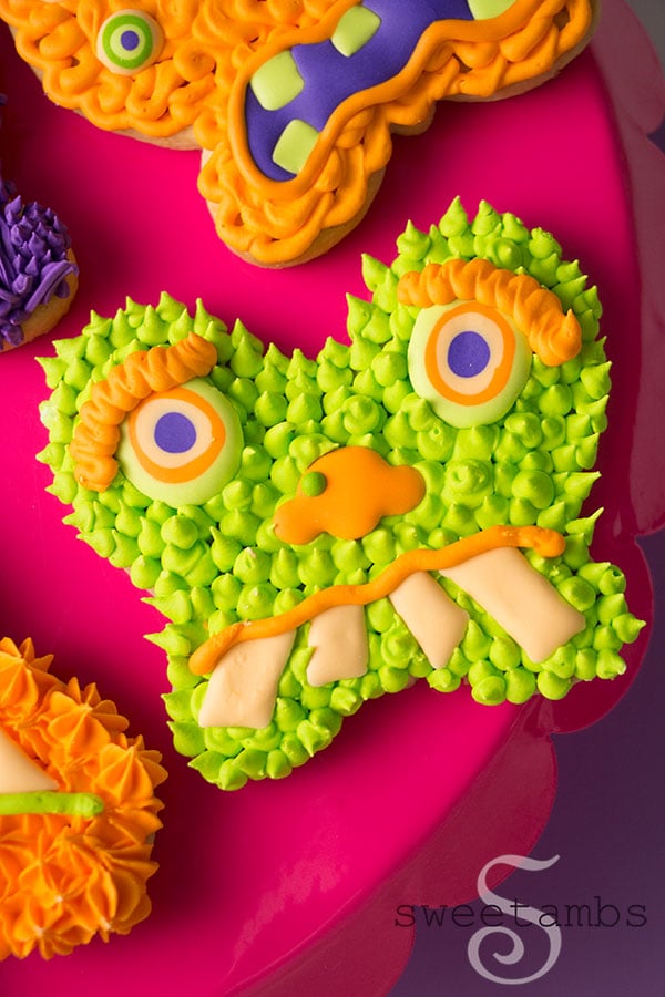 A close-up of Monster Cookies for Halloween - these cookies are cut from a butterfly cookie cutter and decorated to look like monster faces. The monsters are decorated in shades of green, orange, and purple royal icing with lots of different textures. The cookies are on a bright pink cake plate on a purple background.