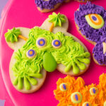 A close-up of Monster Cookies for Halloween - these cookies are cut from a butterfly cookie cutter and decorated to look like monster faces. The monsters are decorated in shades of green, orange, and purple royal icing with lots of different textures. The cookies are on a bright pink cake plate on a purple background.