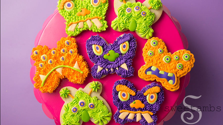 Monster Cookies for Halloween - these cookies are cut from a butterfly cookie cutter and decorated to look like monster faces. The monsters are decorated in shades of green, orange, and purple royal icing with lots of different textures. The cookies are on a bright pink cake plate on a purple background.