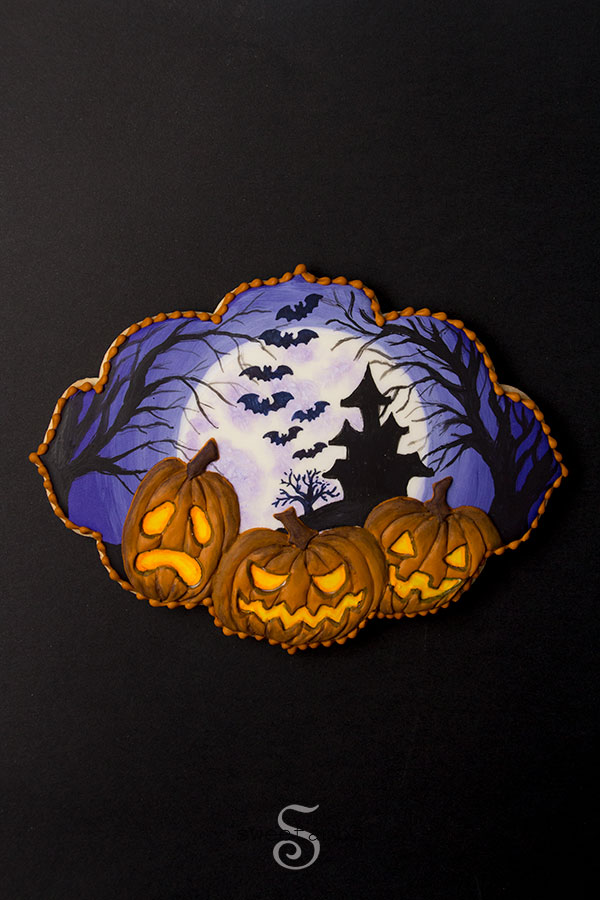 Spooky Jack O'Lantern Cookie for Halloween - a plaque shaped cookie decorated with a full moon, a silhouette of a haunted house on a hill surrounded by spooky trees and bats. 3 glowing jack o'lanterns are in the foreground. There is an orange bead border around the edge of the cookie.