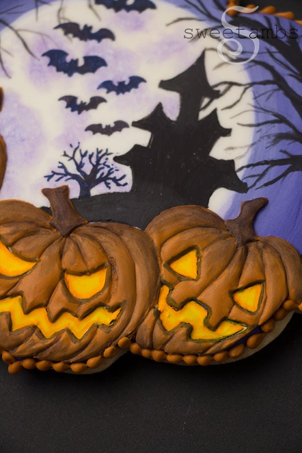A close up of the Spooky Jack O'Lantern Cookie for Halloween - a plaque shaped cookie decorated with a full moon, a silhouette of a haunted house on a hill surrounded by spooky trees and bats. 2 glowing jack o'lanterns are in the foreground. There is an orange bead border around the edge of the cookie.