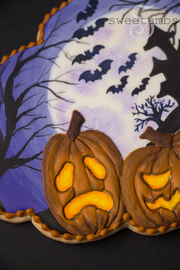 A close up of the Spooky Jack O'Lantern Cookie for Halloween - a plaque shaped cookie decorated with a full moon, a silhouette of a haunted house on a hill surrounded by spooky trees and bats. 2 glowing jack o'lanterns are in the foreground. There is an orange bead border around the edge of the cookie.