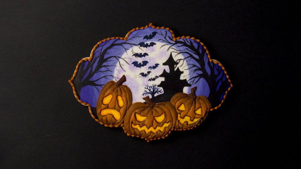 Spooky Jack O'Lantern Cookie for Halloween - a plaque shaped cookie decorated with a full moon, a silhouette of a haunted house on a hill surrounded by spooky trees and bats. 3 glowing jack o'lanterns are in the foreground. There is an orange bead border around the edge of the cookie. 